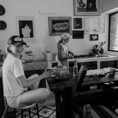 Two people in the Parco dei Sesi kitchen — one laughing on a stool, one cooking at the stove, gallery wall behind